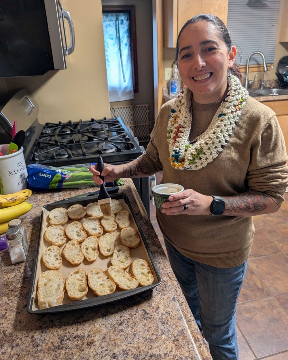 Amy smiling in a home kitchen, brushing a tray of sliced baguette for crostini